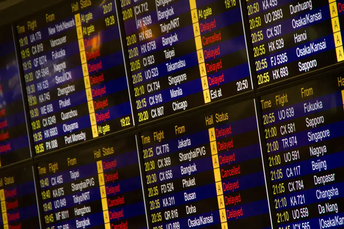 Passengers sitting in airport terminal with luggage, flight information displays showing cancellations, concerned expressions, airport seating area