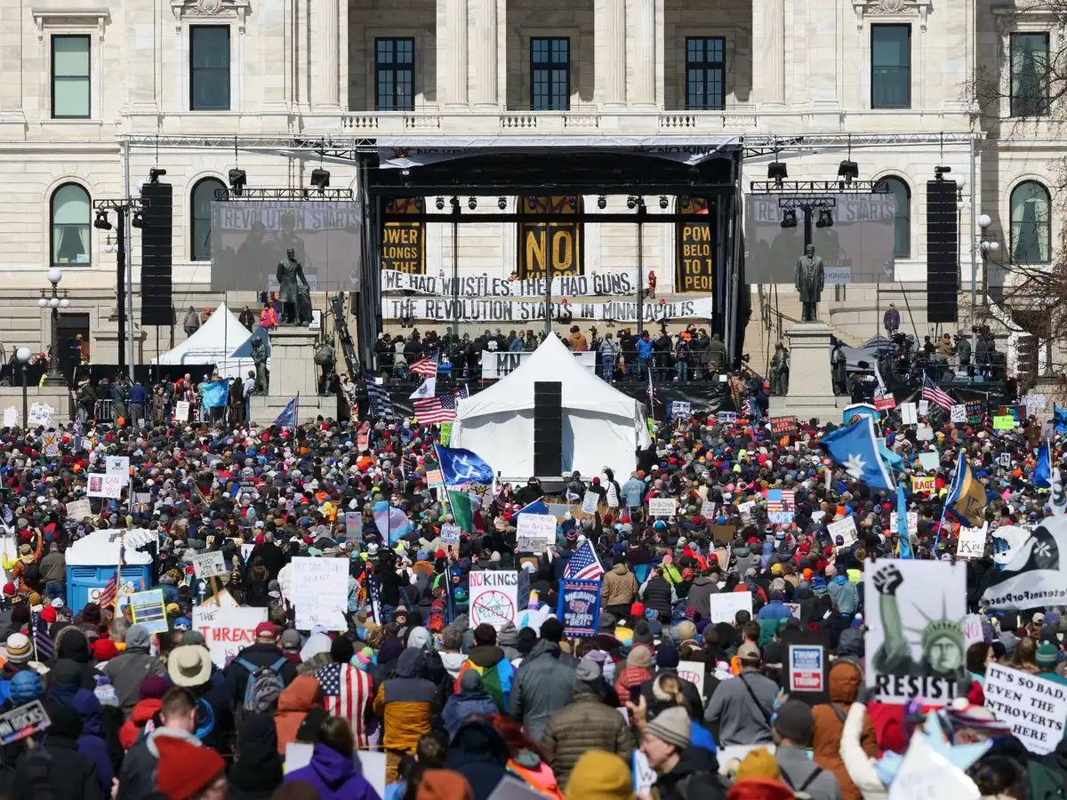 Campaign rally stage with podium and microphones under bright lights, anonymous crowd silhouettes in motion, energy and anticipation visible in the background