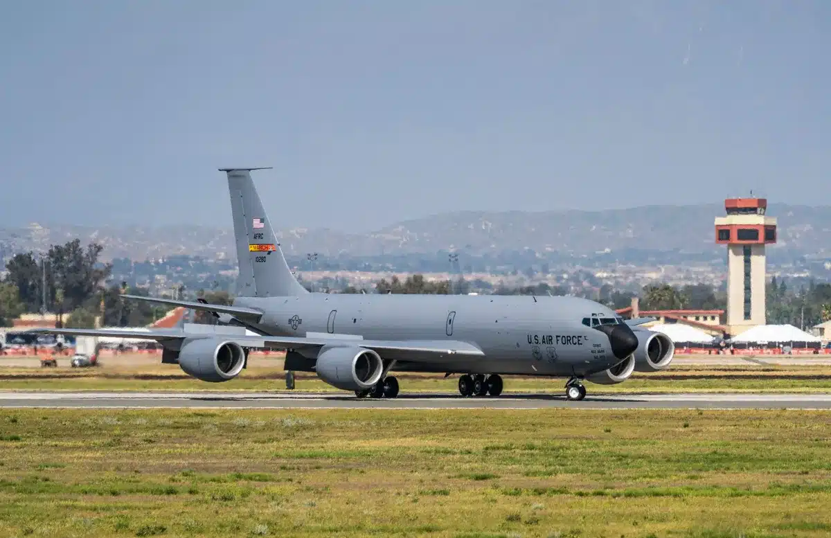 An older Air Force One aircraft on a tarmac at an air base, with ground crew visible nearby, military hangars in soft focus behind it.