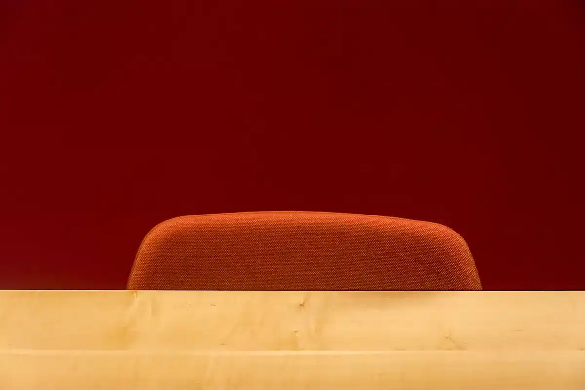 A television studio set with empty anchor desk, single spotlight illuminating the chair, control room lights blurred in the background, papers scattered on the desk, a sense of production paused