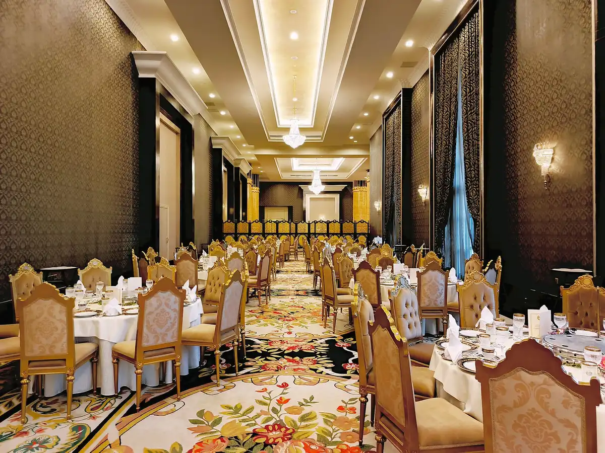 A grand ballroom decorated with formal place settings and centerpieces, chandeliers glowing overhead, empty chairs arranged for an elegant dinner, security barriers visible in soft focus