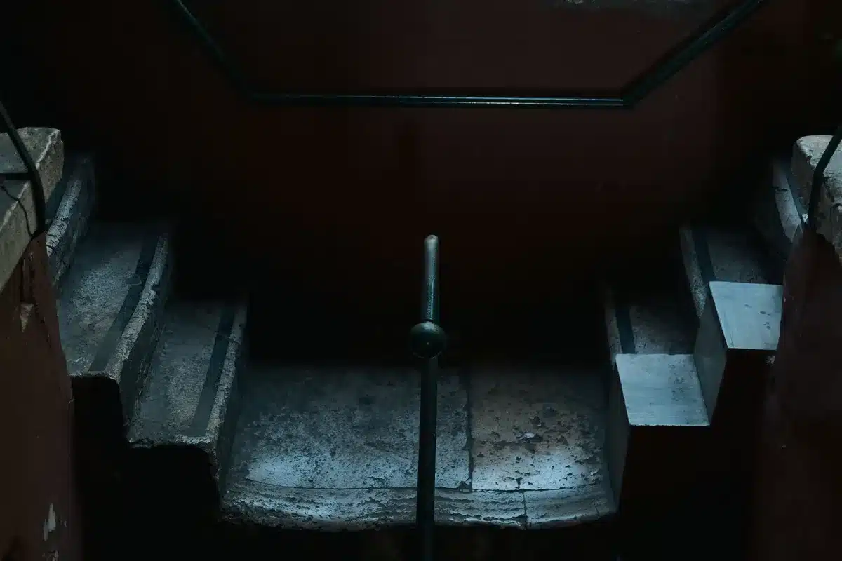 An 80-year-old man navigating a steep metal staircase with visible uncertainty, gripping the railing firmly, one measured step at a time, fluorescent overhead lighting casting sharp shadows, the stairwell empty and clinical