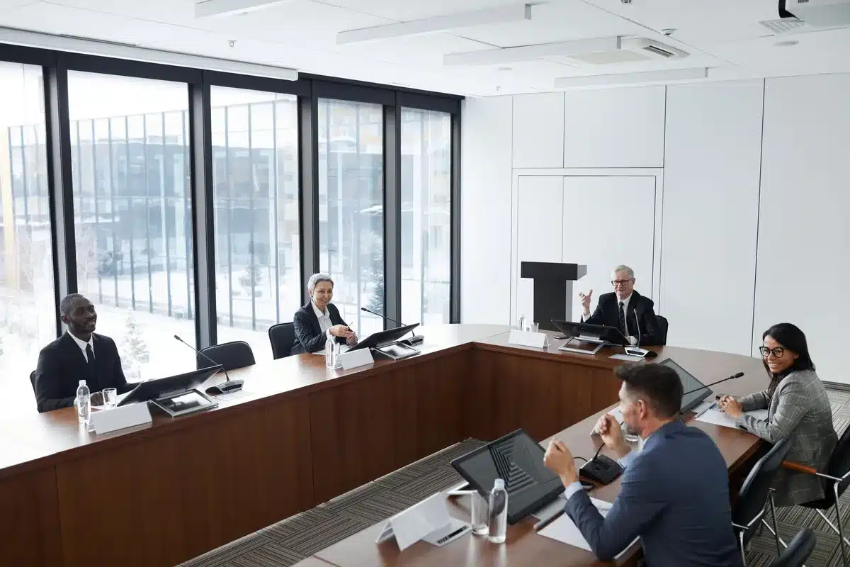 Cabinet officials seated around a polished table in a formal meeting room, papers and water glasses arranged before them, afternoon light streaming through tall windows, serious discussion underway