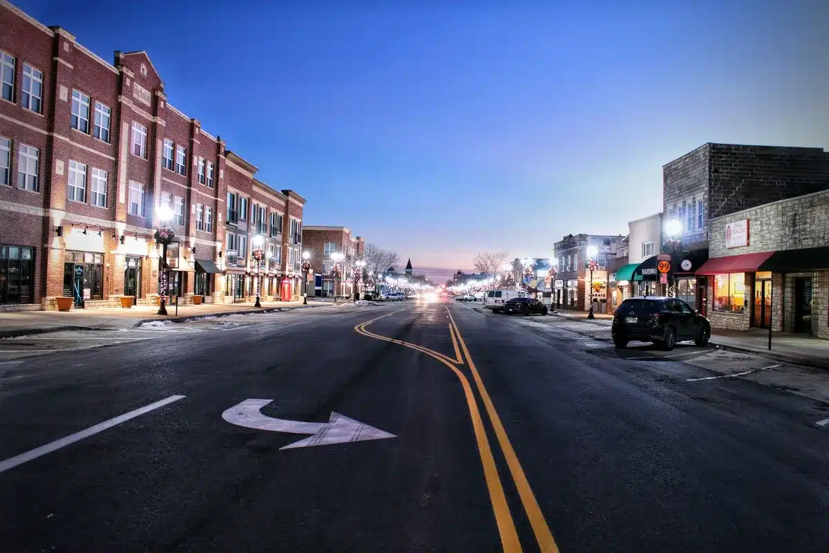 Rural town main street at dusk, storefronts and church spire silhouetted, a few vehicles parked along quiet street