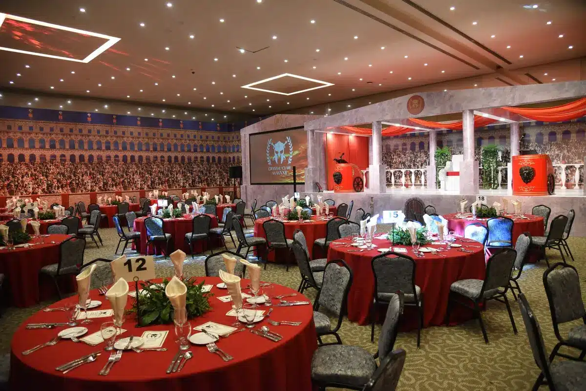 A formal ballroom prepared for an elegant dinner event, with tables set with white linens, place settings, and soft lighting, security barriers visible at entry points, no people present