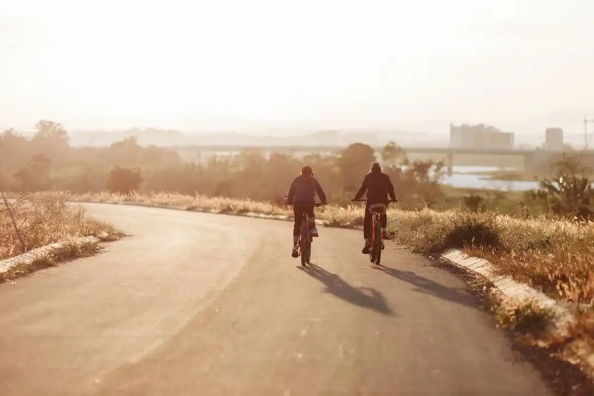 A handcyclist in motion on an open rural road, hands propelling a three-wheeled hand cycle forward, rolling through Iowa farmland under summer sunlight, dust or gravel visible beneath the wheels, determination in the forward motion, small town buildings visible in the distance