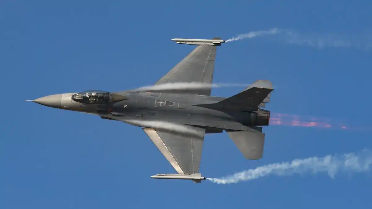 A Ukrainian F-16 fighter jet in flight with air-to-air missiles mounted, silhouetted against a cloudy sky over a vast landscape.