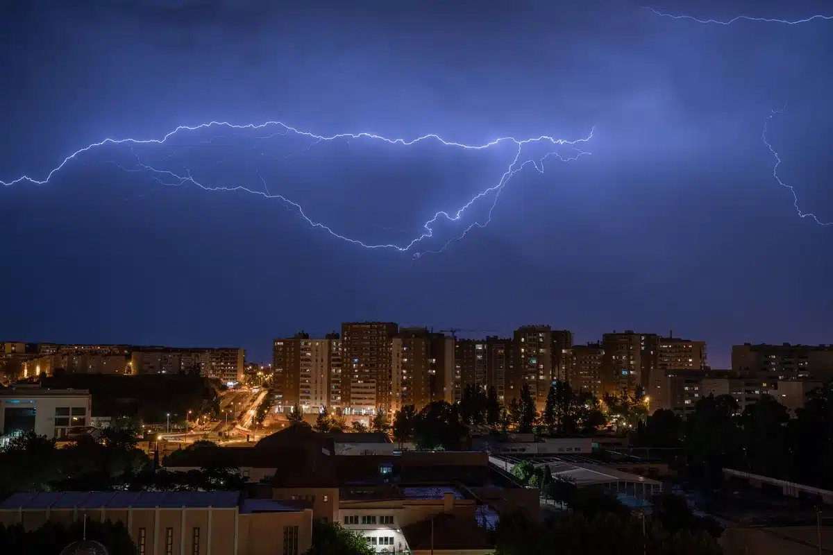 A storm system moving across an urban landscape with dark clouds overhead and lightning visible, contrasting with parched earth and cracked ground in the foreground, showing the stark shift between weather extremes.