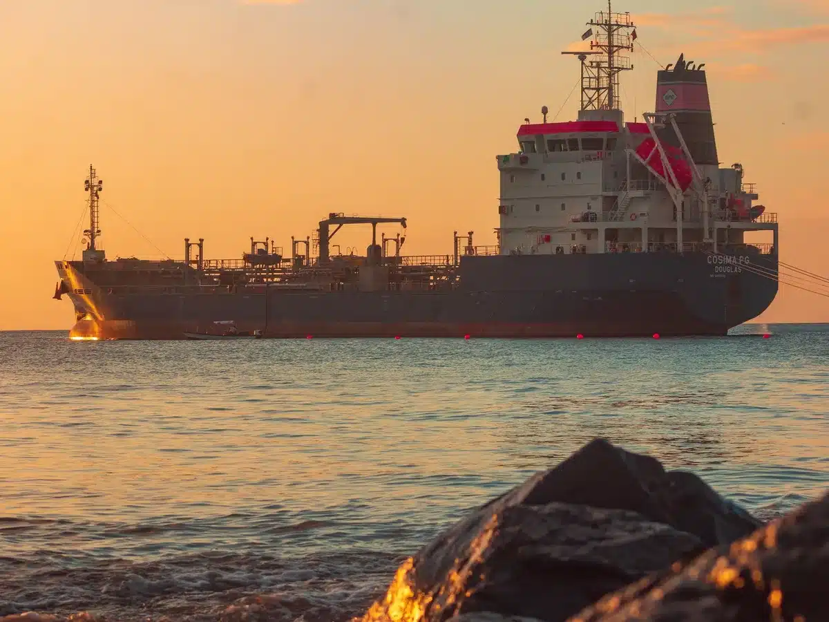 Oil tanker navigating through narrow strait waters with military vessels in distant background, geopolitical tension visible