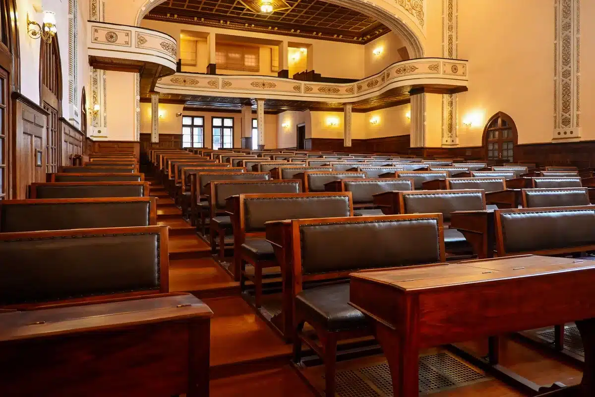 A modern legislative chamber or meeting hall with empty seats arranged in rows, soft overhead lighting illuminating the space, a few scattered papers on desks, a sense of quiet anticipation.