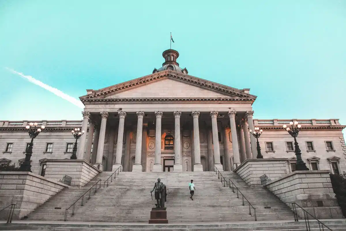 Capitol building steps at dawn or dusk, empty and austere, American flag in background, suggesting the site of political protest and official scrutiny.