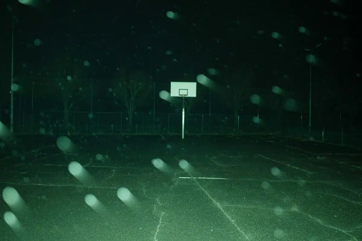 An empty basketball arena court at night, shadows cast by overhead lights, a single player's silhouette visible in the distance, the scoreboard dark except for the final score glowing, the court markings sharp and clear