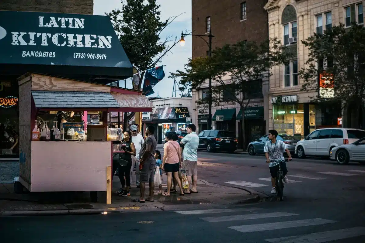 A crowded outdoor street festival scene with colorful vendor booths, string lights, and anonymous fans in casual attire mingling and holding drinks, neon signage glowing in the background, vibrant energy and celebration