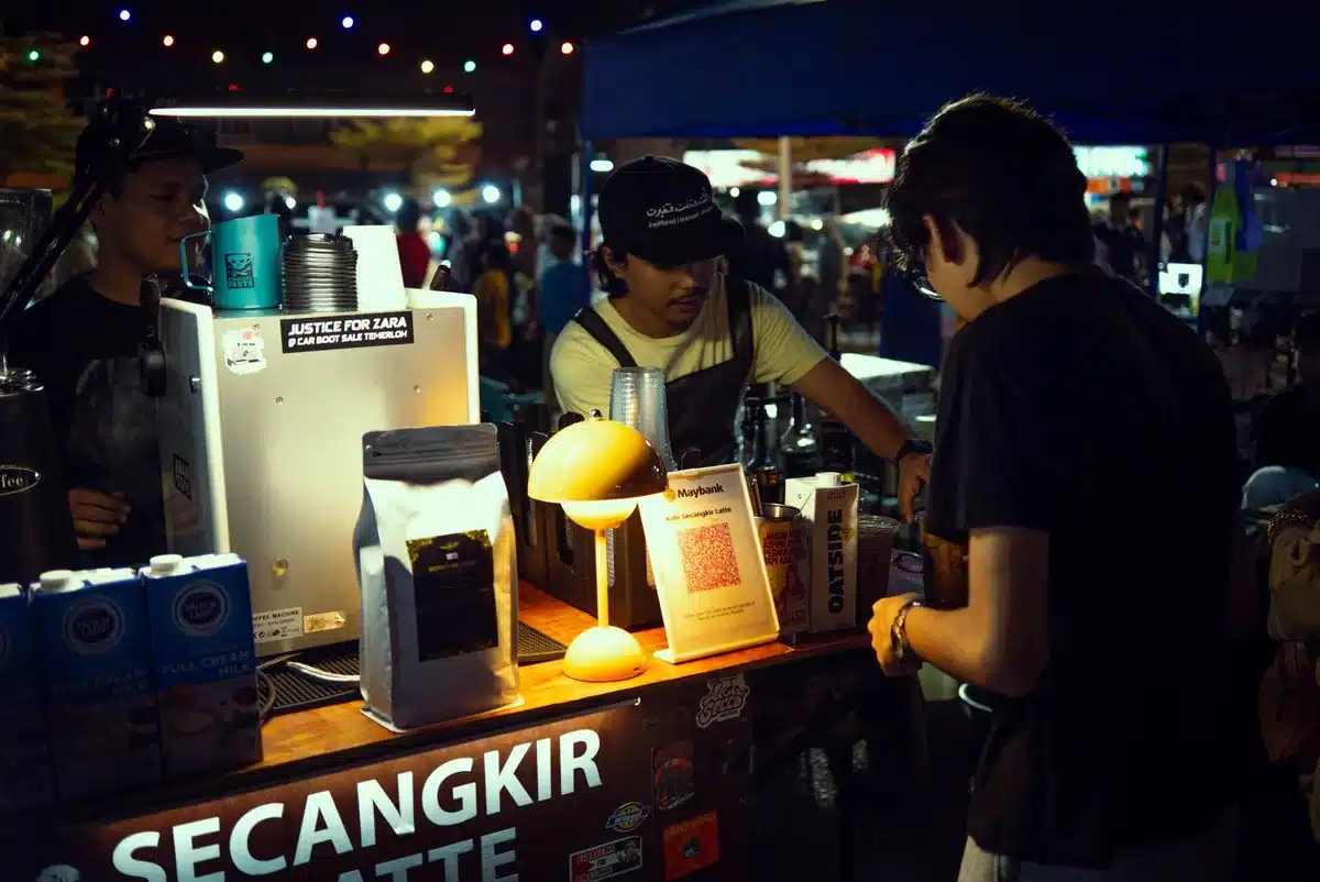 A vendor booth display at a festival with Unwell Hydration branded bottles and merchandise arranged on a table, attendees reaching for products, daylight streaming across the setup