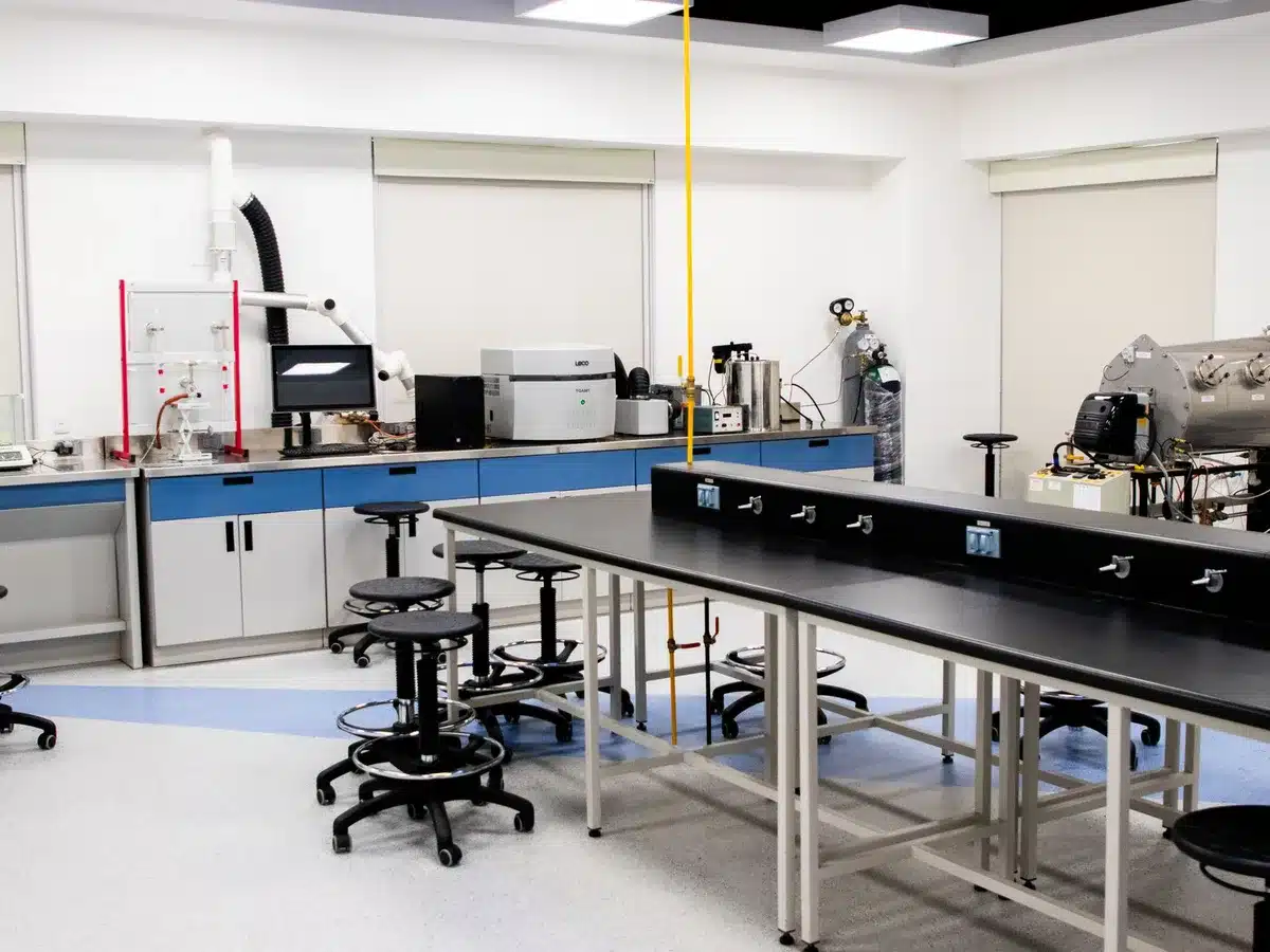 An empty AI research laboratory workspace with computer screens displaying code and neural network visualizations, fluorescent lighting casting a clinical atmosphere, a single desk chair in shadow