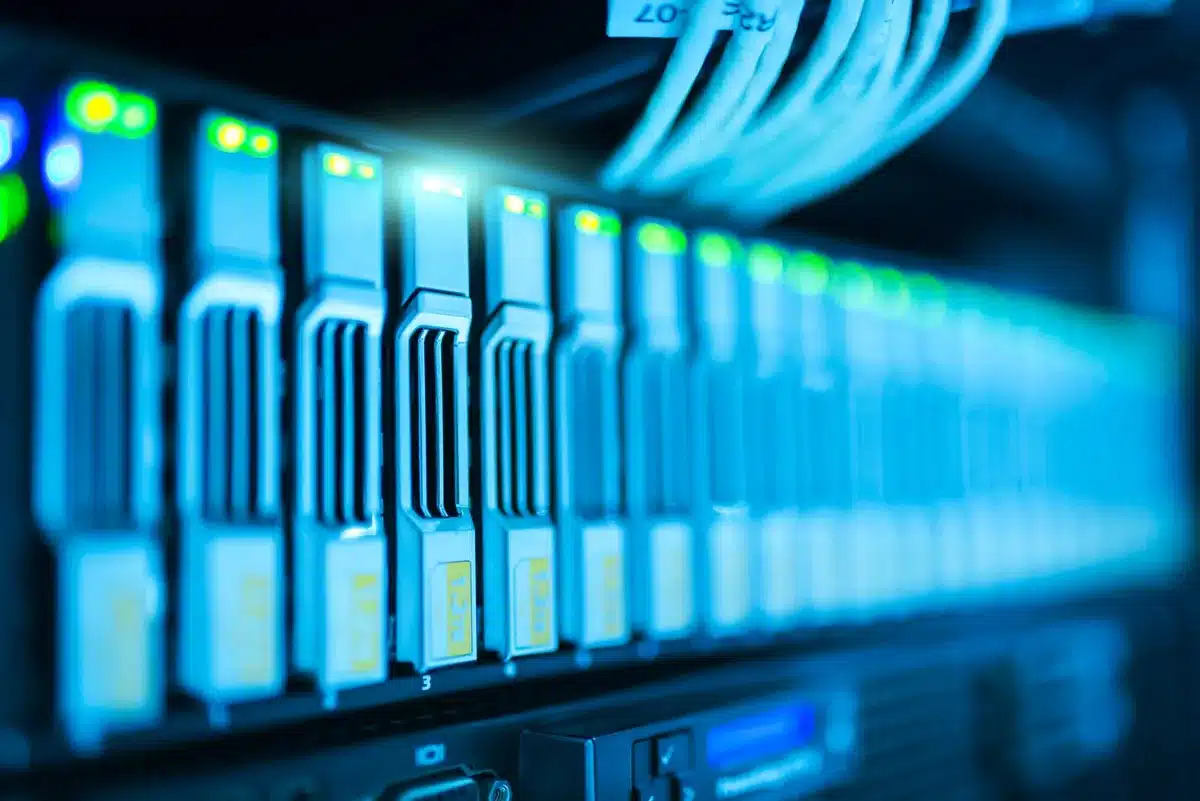 Server racks with blue and green indicator lights illuminating in a vast data center, rows extending into shadow, ambient glow reflecting off metal surfaces