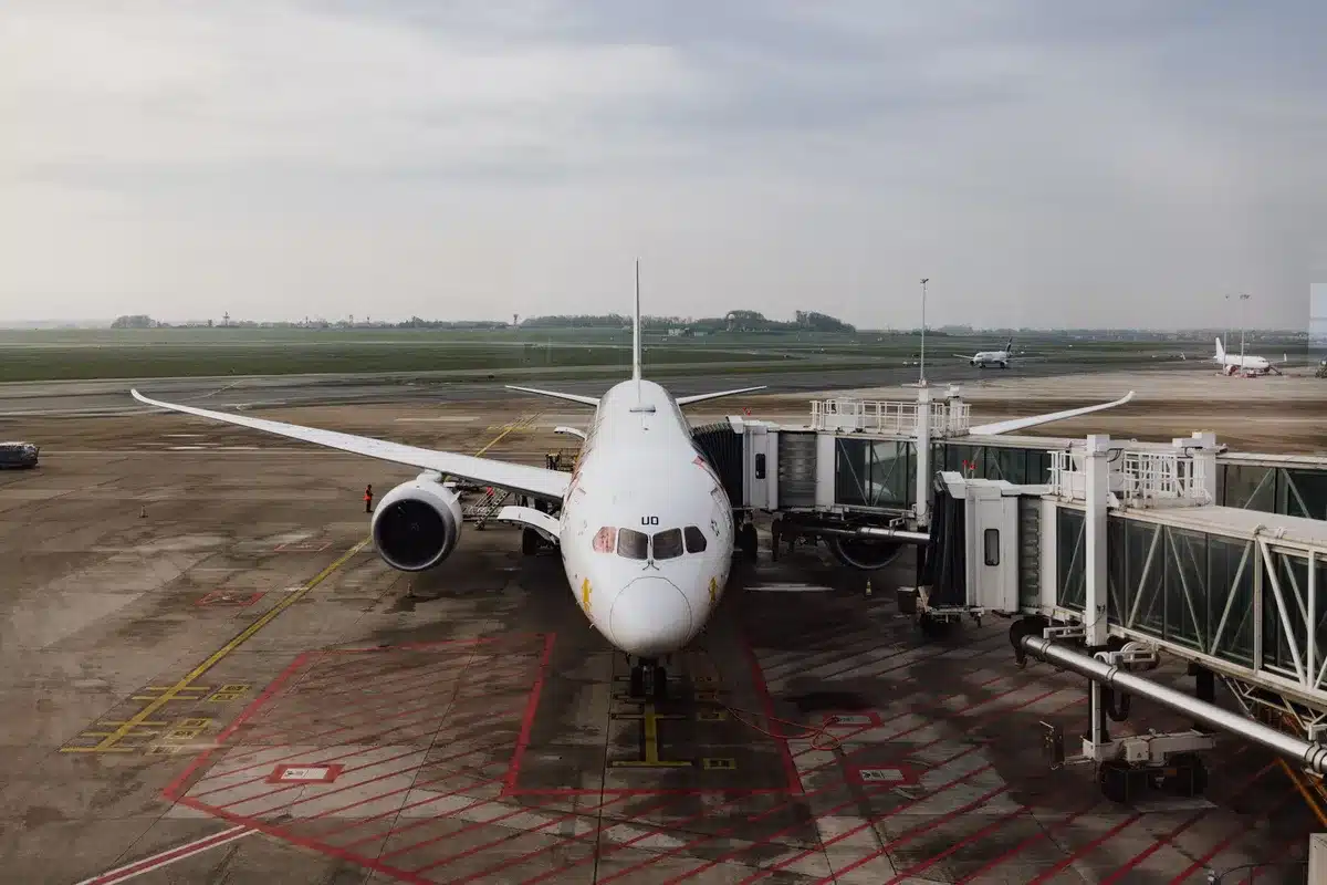 Wide-body aircraft on tarmac at sunset with ground crew and ground support vehicles, European airport signage visible in background.