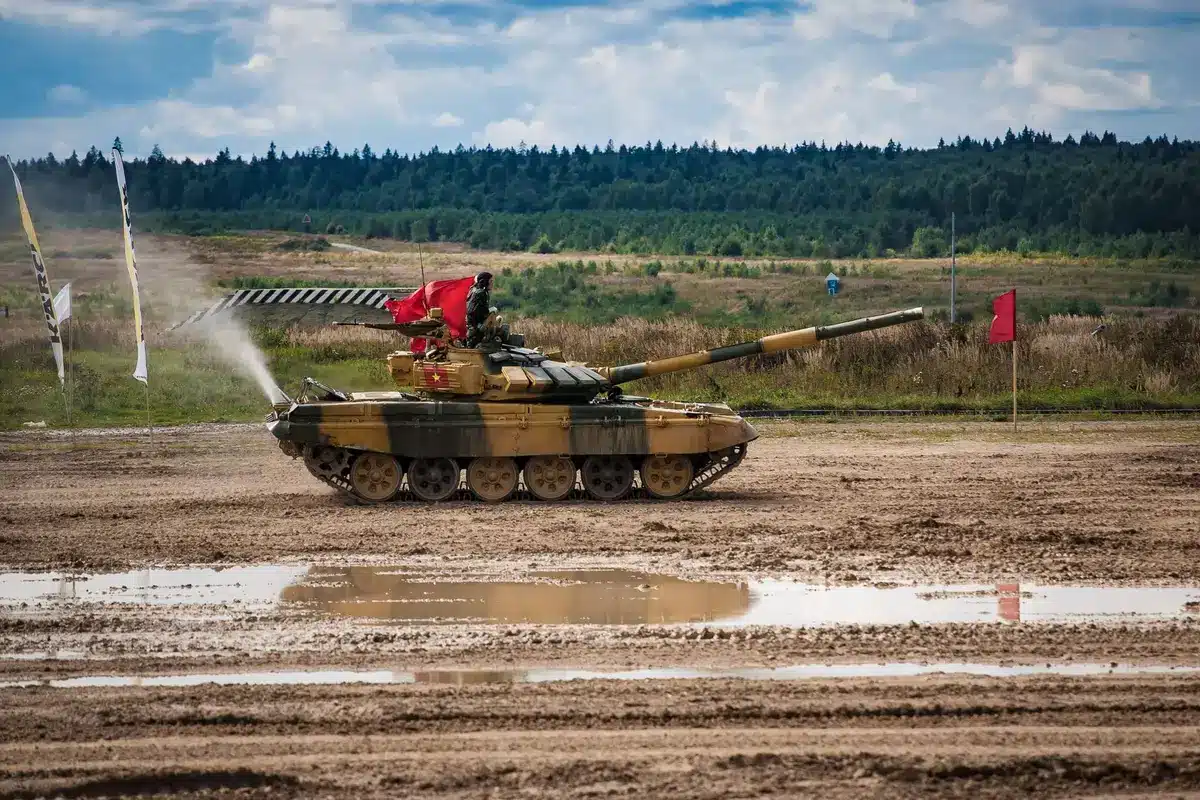 A man in camouflage stands beside a Soviet-era military tank on an open training ground, weapons visible nearby, amid a battlefield-style environment under overcast sky