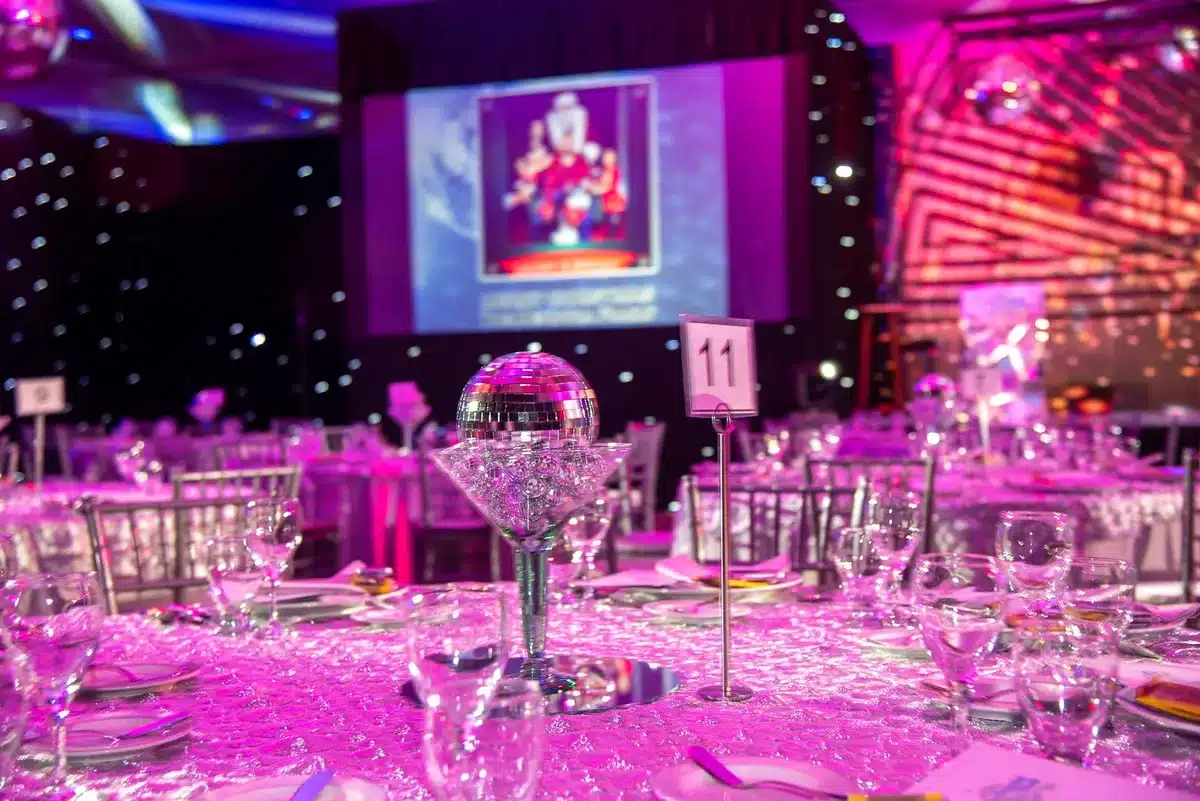 A formal gala banquet hall with rows of elegant place settings and centerpieces, a stage with microphone and podium in soft focus, anonymous attendees in formal wear seated at round tables in warm ambient lighting
