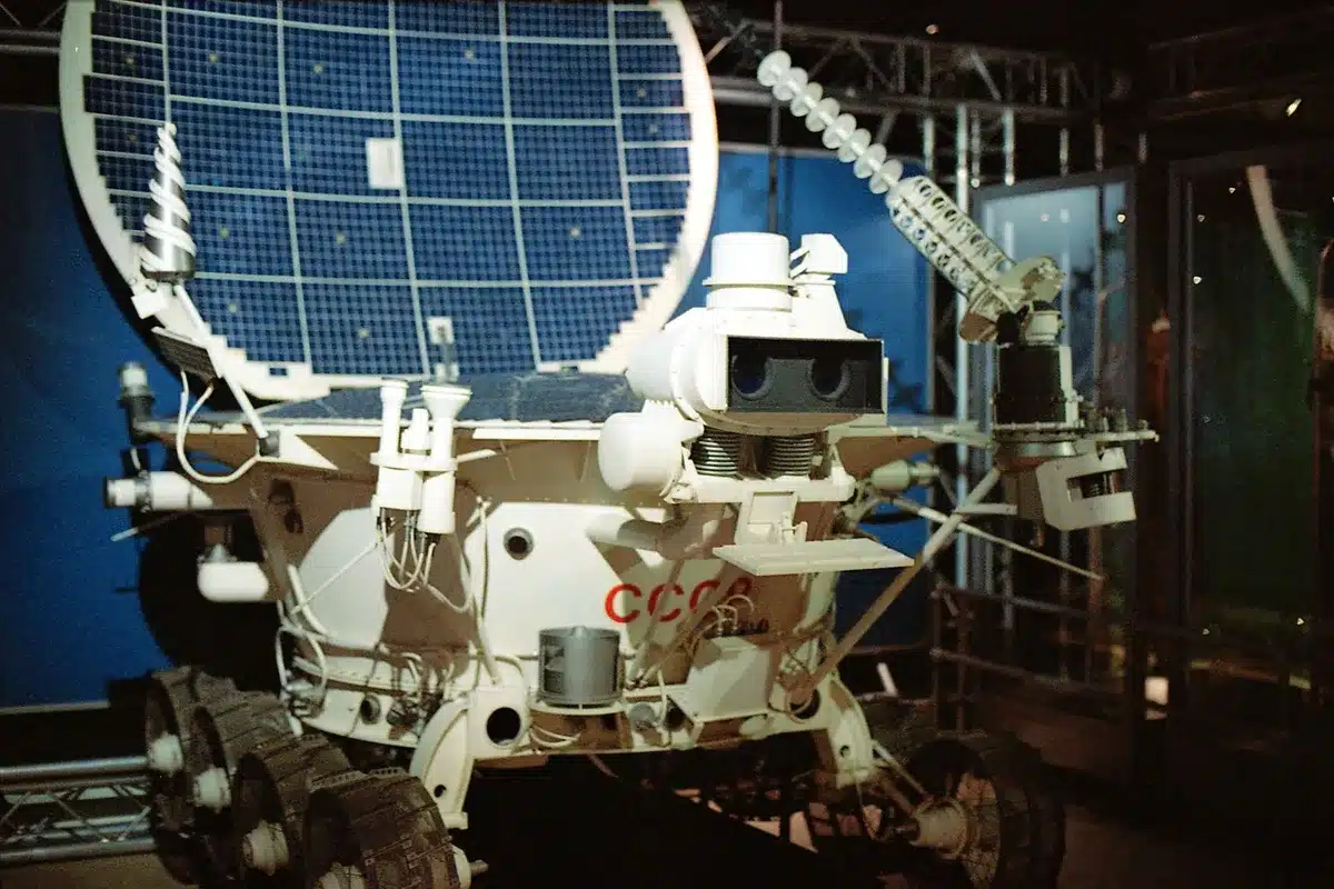 A lunar rover silhouette against a stark moon horizon, with distant mountains in the background, wheels kicking up dust as it travels across gray regolith.