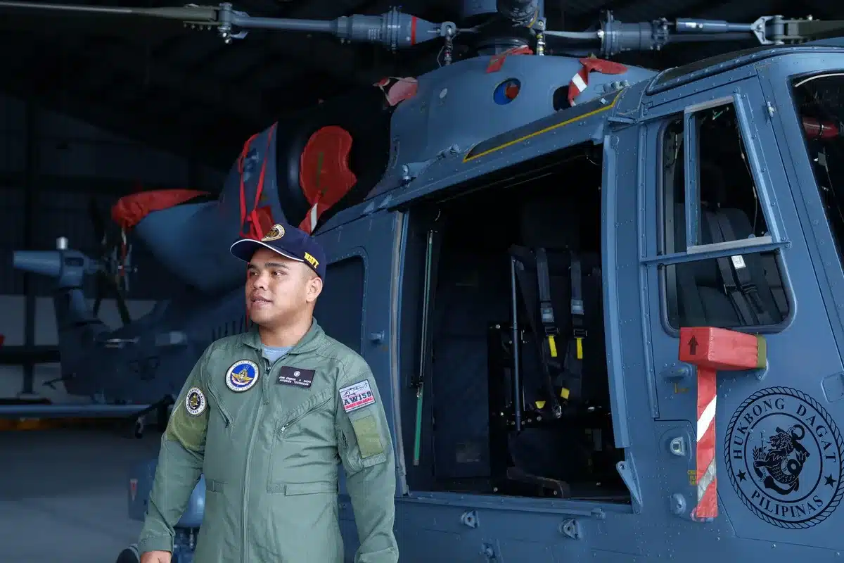 Two soldiers in military gear standing in front of an AH-64 attack helicopter on a tarmac, sunlight catching the aircraft's distinctive twin rotors and nose turret, a sense of routine readiness.