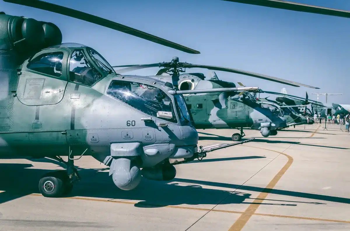 A vast military airfield at dawn with multiple AH-64 Apache helicopters lined up in rows on the tarmac, rotors still, no personnel visible, a sense of operational pause.