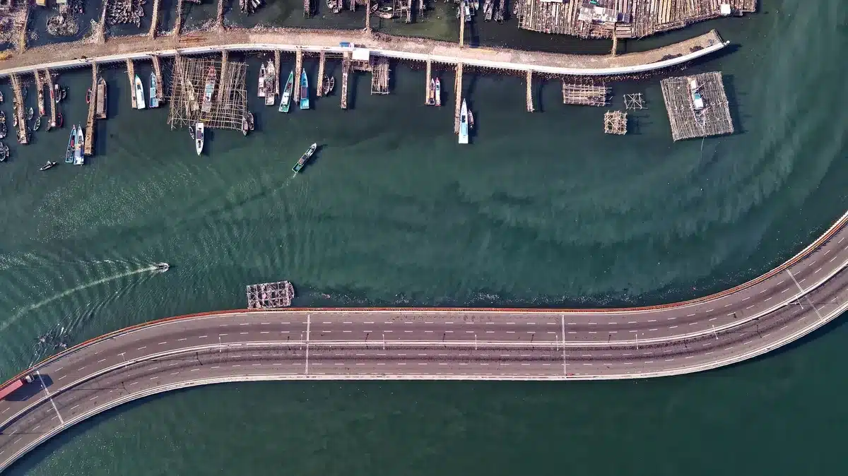 Satellite view of a newly constructed artificial island with geometric seawalls, internal roads, and a lagoon, showing dredging equipment and construction activity in the surrounding waters, surrounded by turquoise ocean.