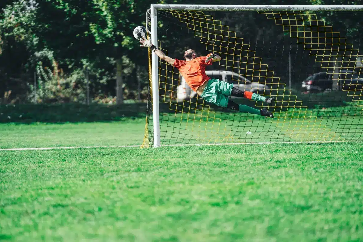 A close-up of a goalmouth scramble, the ball nestled in the net, grass and white netting in sharp focus, a blurred defender in the background.