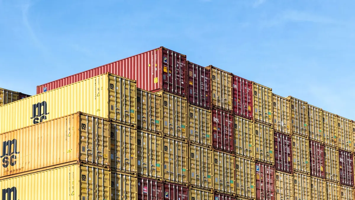 Stacked shipping containers and industrial tariff-related goods at a port or warehouse, with documents and calculator showing cost calculations
