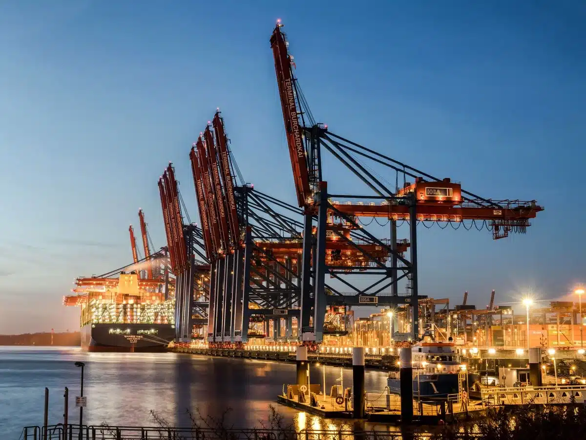 A darkened shipping port at dusk with container cranes silhouetted against a fading sky, a single cargo vessel moored at the dock, no flags or markings visible, fog rolling across the water