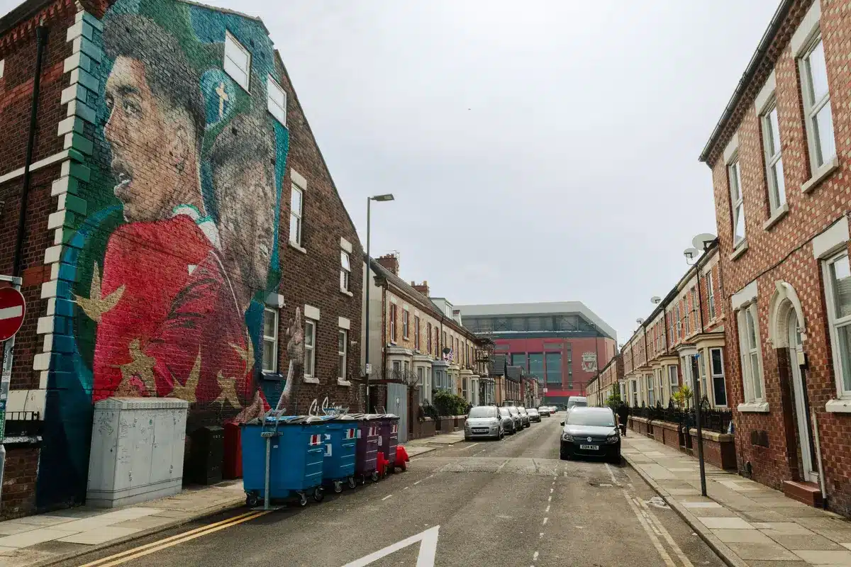 Liverpool Football Club stadium exterior at dusk with floodlights illuminated, the Shankly Gates entrance visible, empty streets suggesting pre-season period