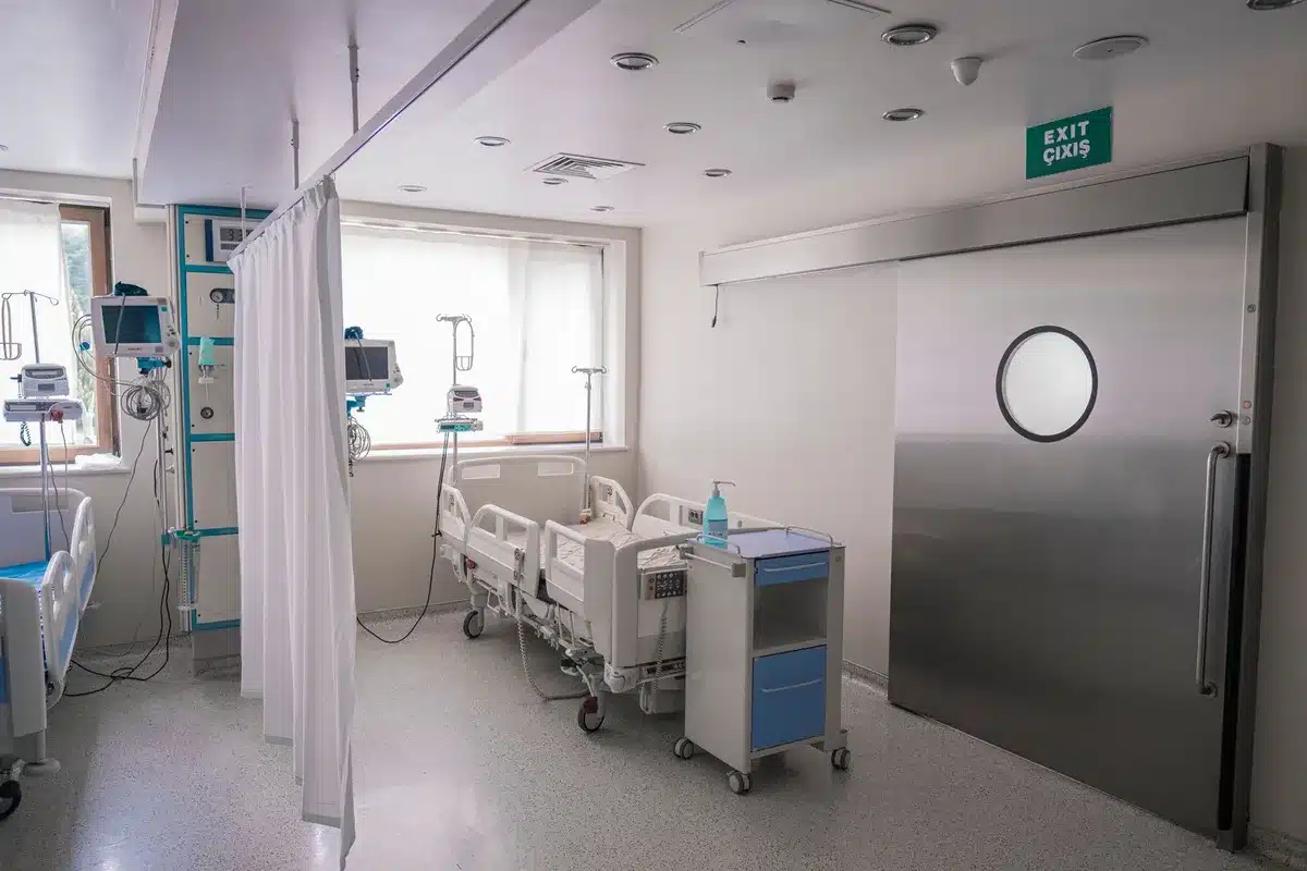 A dimly lit hospital corridor with medical equipment visible but blurred, a single empty chair in shadow, soft blue-tinted lighting suggesting a medical facility in the evening hours