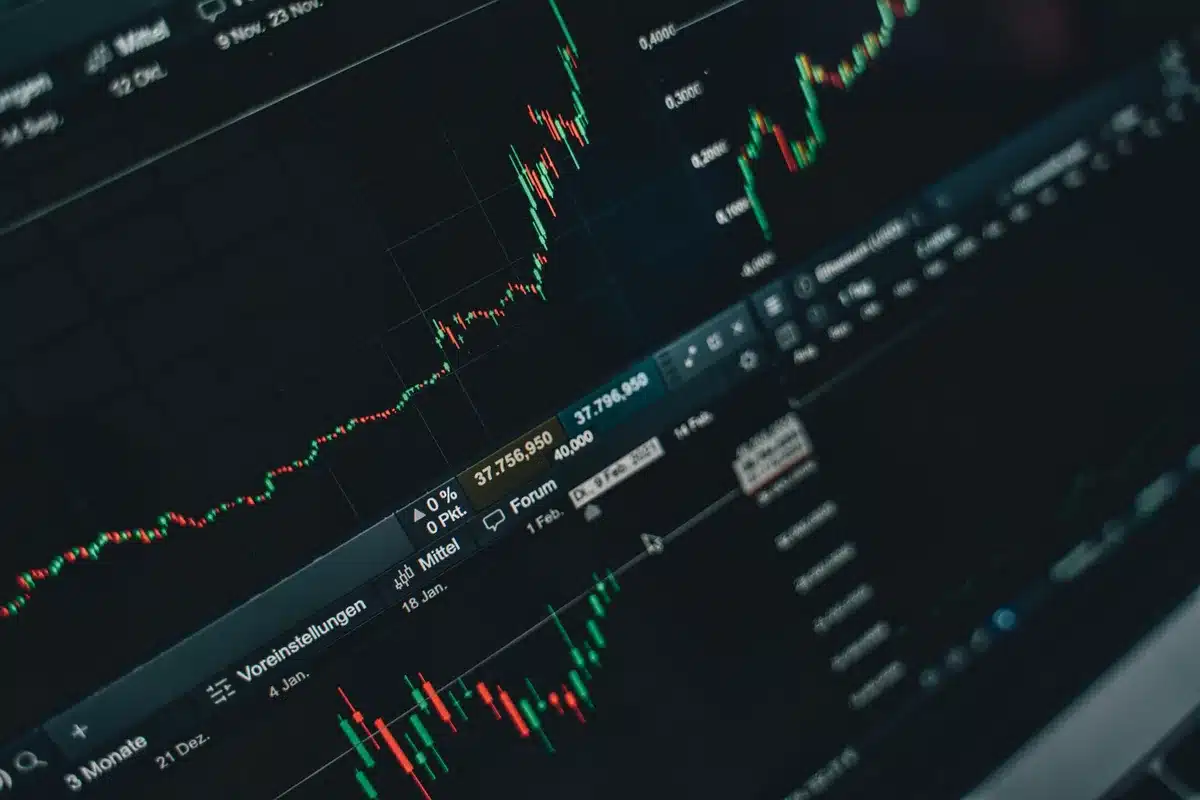 A laptop screen displaying cryptocurrency trading charts with upward trends in green, a smartphone beside it showing Bitcoin price data, papers with regulatory notes scattered across a desk, warm desk lamp illuminating the workspace.