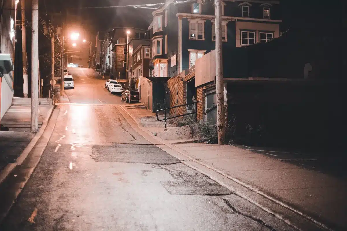 A quiet residential street in Queens at dusk, modest homes with lights on, a single candle or memorial flowers visible on a doorstep, empty sidewalk, somber and still.