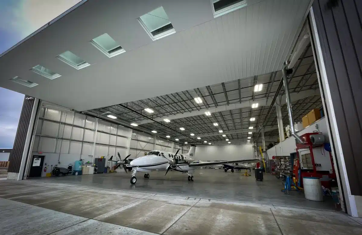 A darkened aircraft hangar with a single 737 fuselage section positioned under bright inspection lights, tools and measurement equipment arranged on a work platform below
