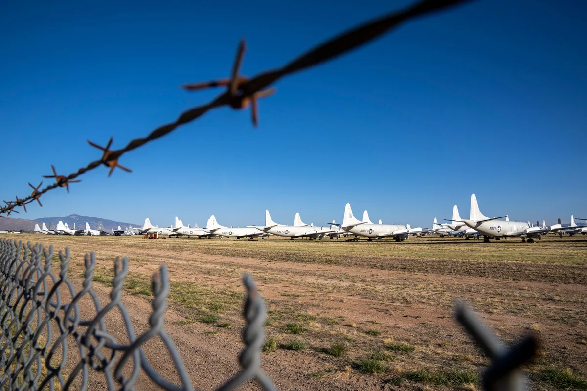 Aircraft fuselage in outdoor storage at an aerospace facility, weathered from years of exposure, surrounded by industrial equipment and desert landscape.