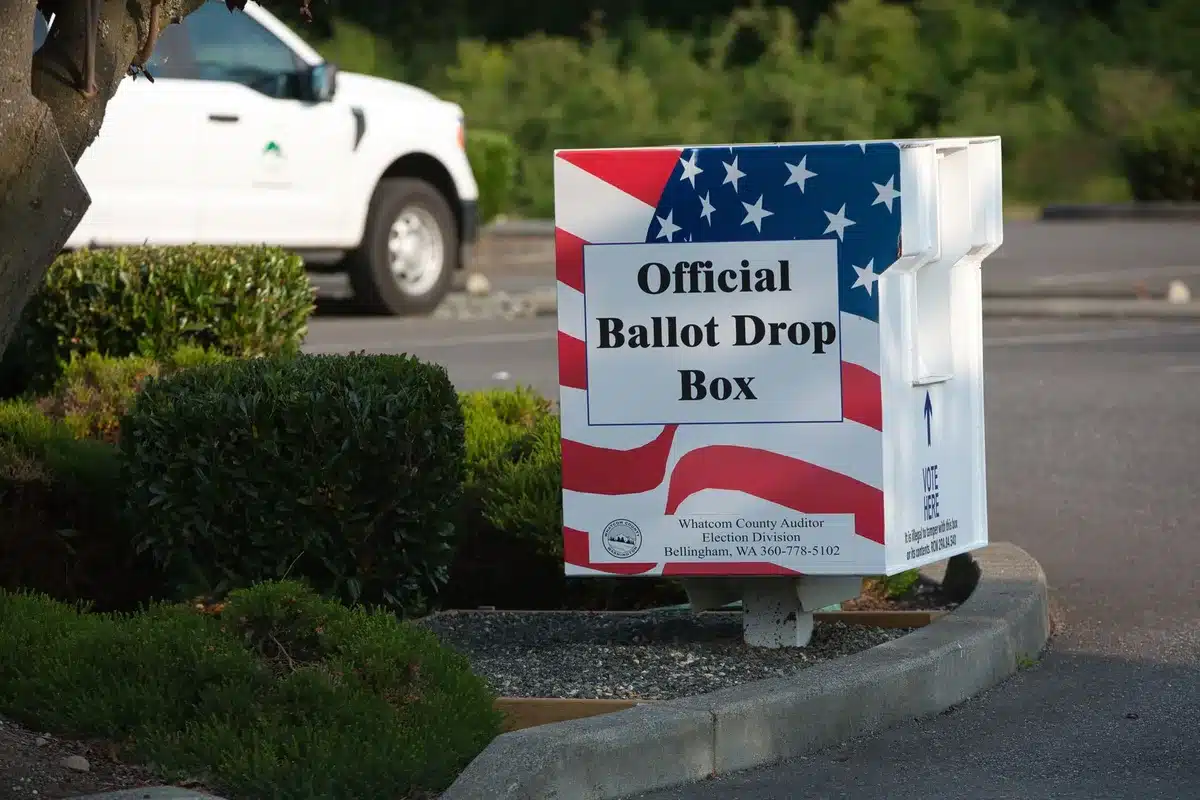 A voting location entrance with directional signs and an American flag, people entering to cast ballots, morning sunlight creating long shadows.