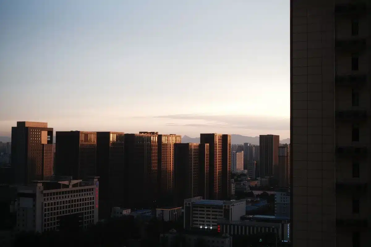 An empty executive office overlooking Omaha skyline at dawn, leather chair facing floor-to-ceiling windows, sunlight breaking through clouds, a framed photograph of the textile mill on the desk&mdash;transition