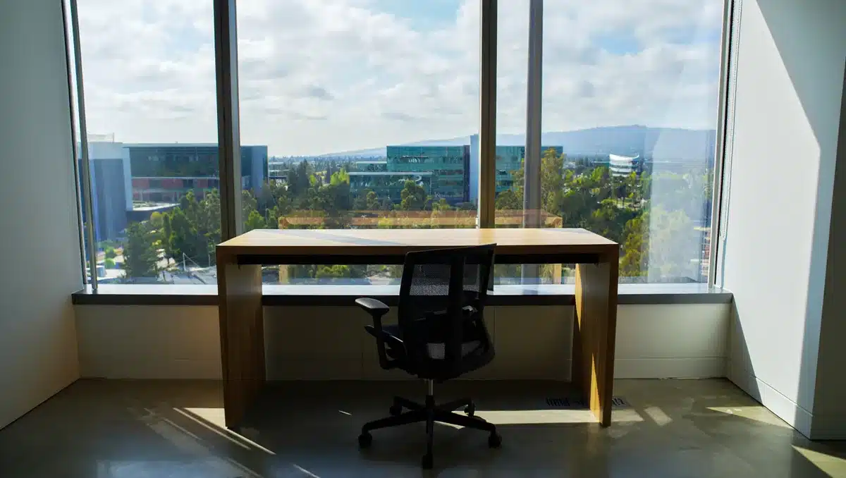 A minimalist office space with large windows overlooking a tech campus, showing an empty desk and chair facing a view of Silicon Valley, suggesting quiet influence behind major decisions