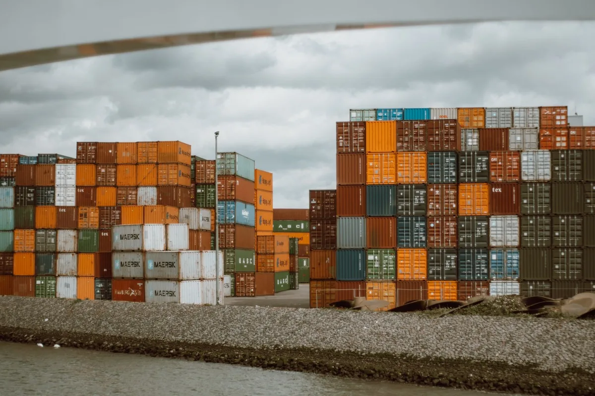 A cargo container at a border crossing, half in shadow, with tariff notice documents visible, a customs officer silhouette in the distance, early morning light suggesting the imminent September 8 start date.