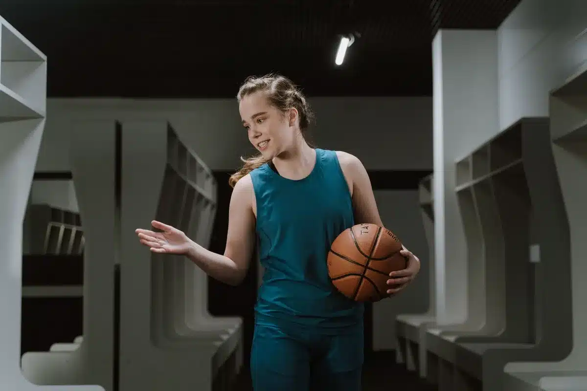 A young basketball player in a Syracuse jersey standing at center court, looking up at the arena lights, teammates visible in soft focus in the background