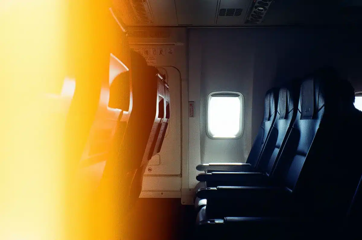 An airplane cabin with window shades drawn, muted overhead lighting, and empty seats suggesting a quiet, uncomfortable moment.