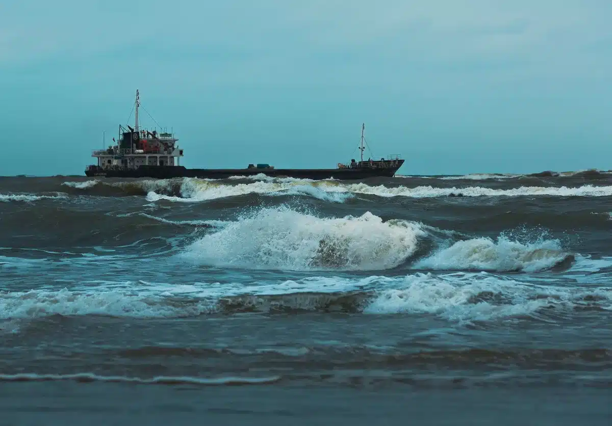 A container ship listing slightly in calm water, its deck laden with stacked cargo containers in fading light, a single tug boat approaching nearby, vulnerability and isolation in the scene