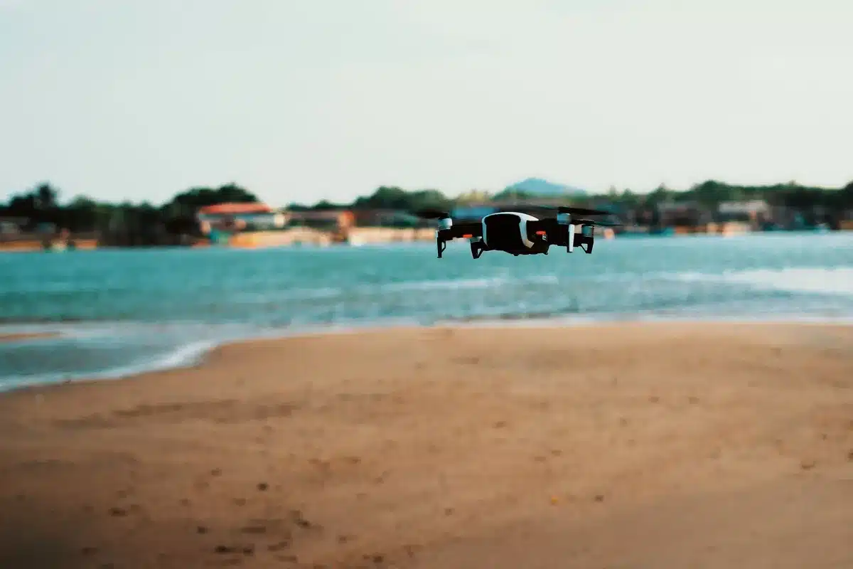 Silhouette of a one-way attack drone suspended mid-air above a blue ocean horizon, backlit by sunset, with the outline of a distant naval vessel below on the water, conveying the multi-domain scope of operations