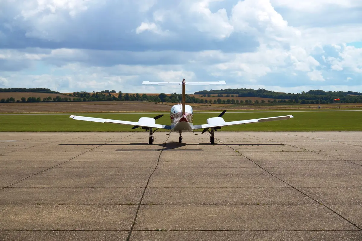 A single-engine Cessna aircraft sits on grass beside a rural highway under clear skies, with utility poles and power lines visible in the distance, and a truck passing on the road behind.