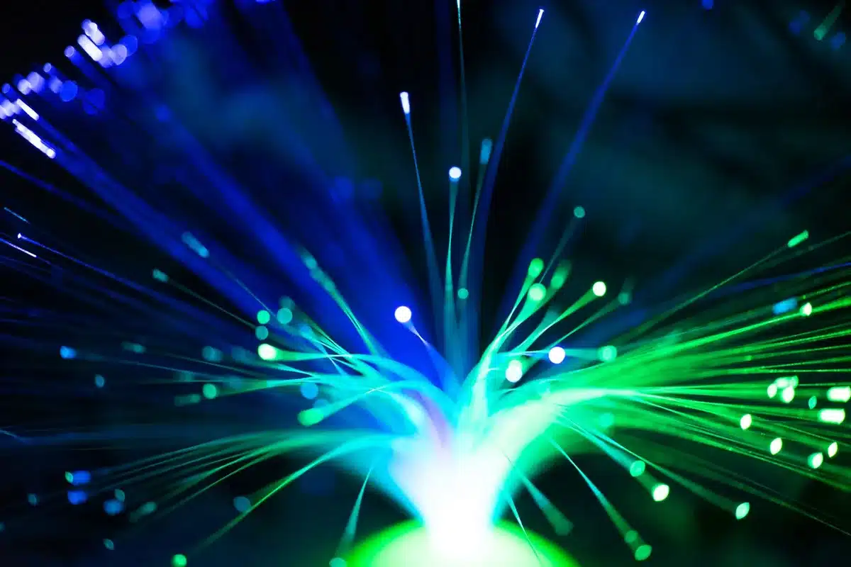 Two anonymous figures examining fiber optic cables and network infrastructure in a modern telecommunications facility, warm overhead lighting casting shadows on the equipment, technical complexity conveyed through cables and panels