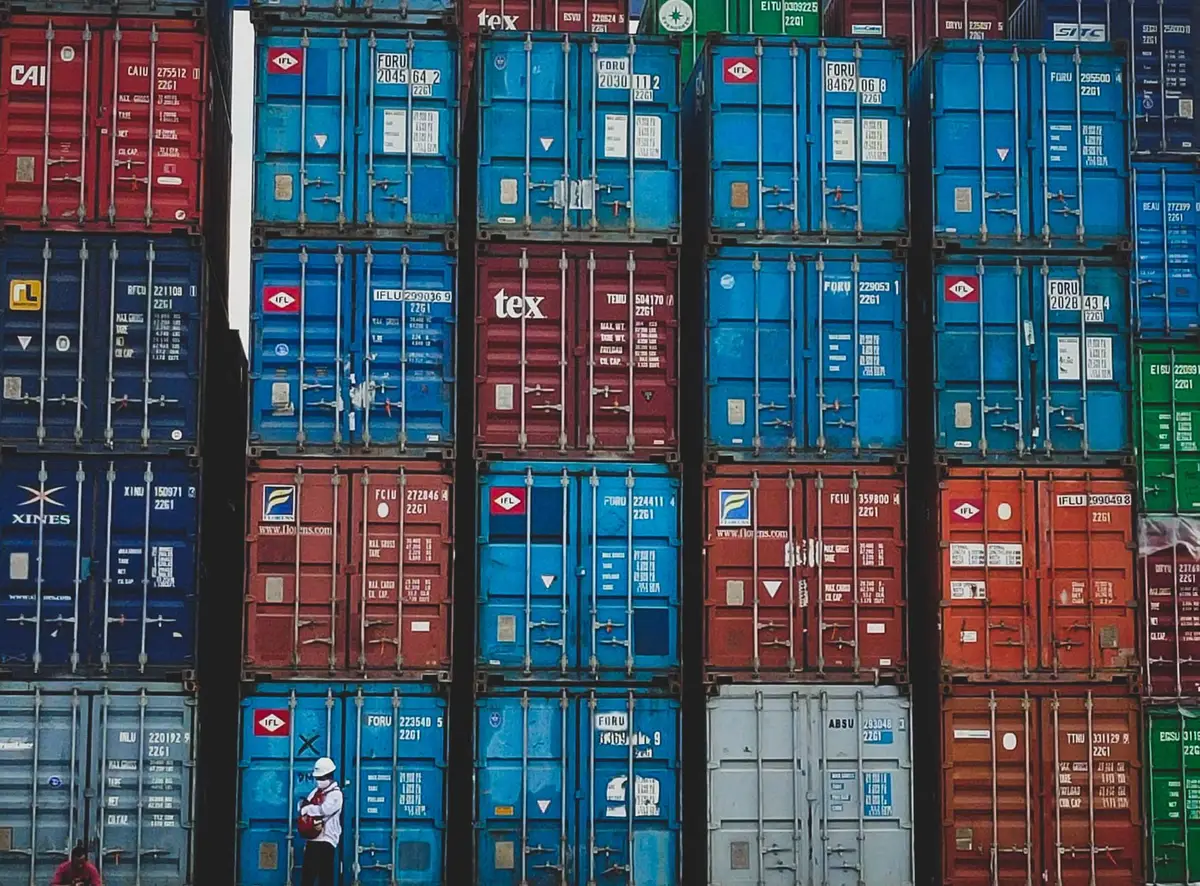 Shipping containers stacked at a busy port terminal, with multiple cargo vessels in the background and industrial cranes loading containers, representing global trade logistics.