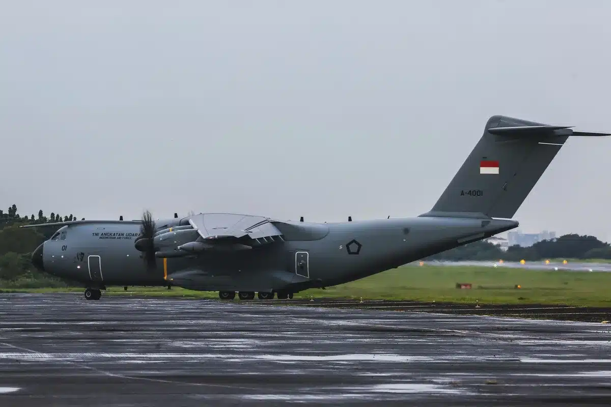 A U.S. military transport aircraft silhouetted against a gray sky, its cargo doors visible, descending toward a distant airport tarmac with Moscow's skyline barely visible through haze.