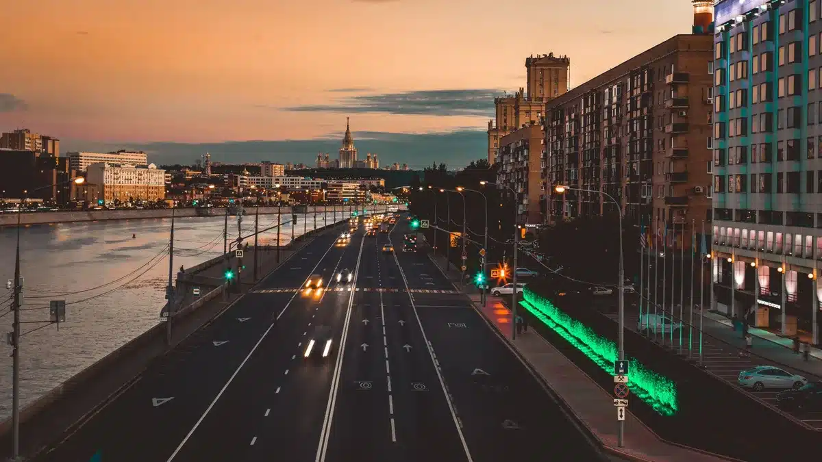 A motorcade of diplomatic vehicles with red license plates driving through a Moscow avenue at dusk, their headlights on, the Kremlin walls visible in shadow on the left.