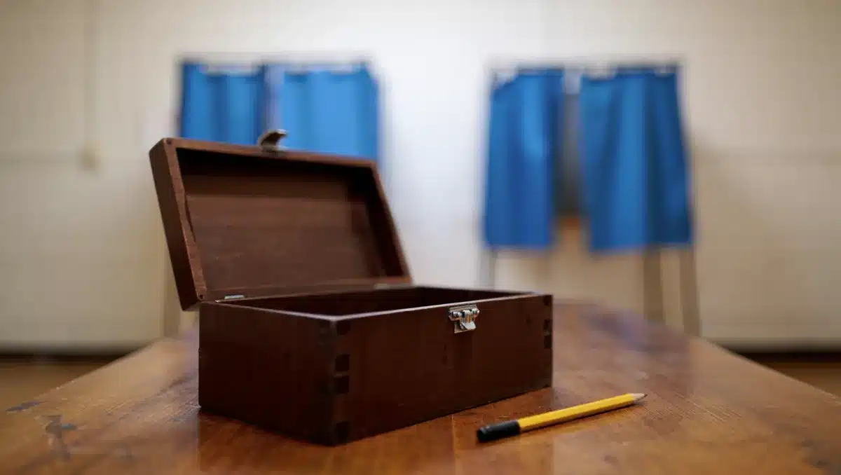An empty Democratic primary ballot box sitting on a wooden table, with voting booths blurred in the background, a single pencil nearby, soft overhead lighting casting subtle shadows &mdash; anticipation.